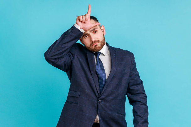 Portrait of upset disappointed young adult businessman with beard in dark suit standing, showing looser gesture with hand on forehead. Indoor studio shot isolated on blue background. - Foto, afbeelding