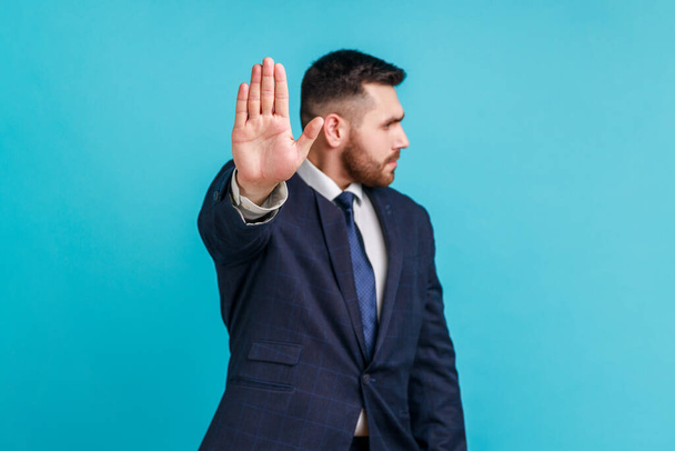 Stop! keep your distance. Serious bearded man wearing official style suit standing with stop hand and looking away, showing ban, block sign with hand. Indoor studio shot isolated on blue background. - Foto, Imagem