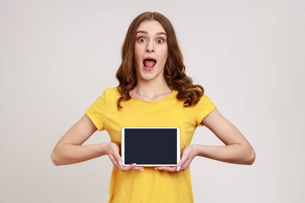 Portrait of shocked beautiful teenage girl in style T-shirt, showing tablet empty screen with unbelievable face, looking at camera with opened mouth. Indoor studio shot isolated on gray background. - 写真・画像