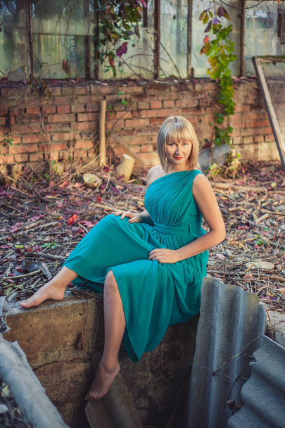 Mujer joven con corte de pelo bob en un vestido de noche de color turquesa en un hombro está sentado en el borde de un hoyo de hormigón en un invernadero abandonado en otoño. Escenario sesión de fotos - basura del vestido - Foto, imagen