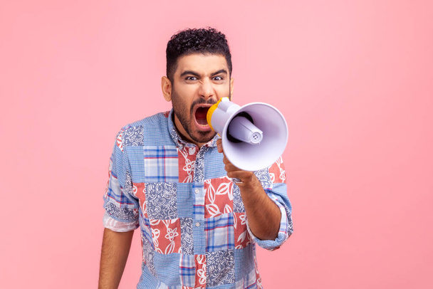 Angry man wearing casual blue shirt holding megaphone near mouth loudly speaking, screaming, making announcement, paying attention at social problems. Indoor studio shot isolated on pink background. - Zdjęcie, obraz