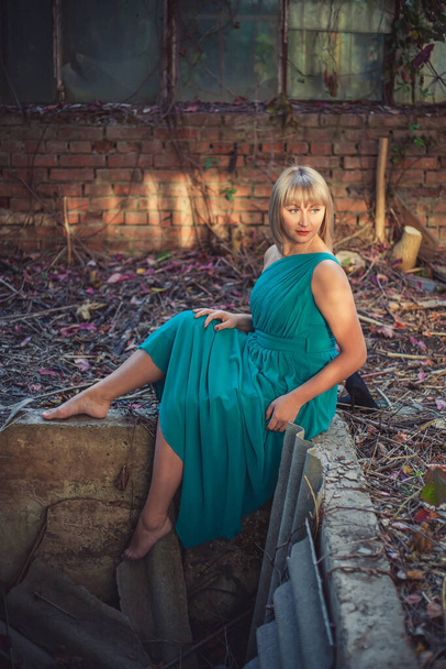 Mladá žena s bob účesem v tyrkysových večerních šatech na rameni sedí na okraji betonové jámy v opuštěném skleníku na podzim. zinscenované focení - smetí šaty - Fotografie, Obrázek