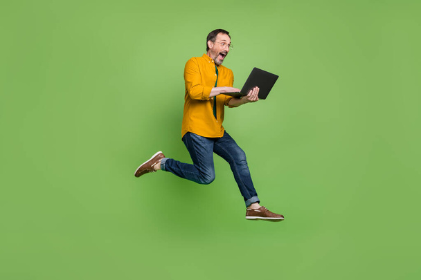 Full size photo of brown haired happy amazed man jump up run hold laptop isolated on green color background - Foto, Bild