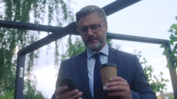 Male boss drinking coffee and texting on phone while walking in park, business - Video