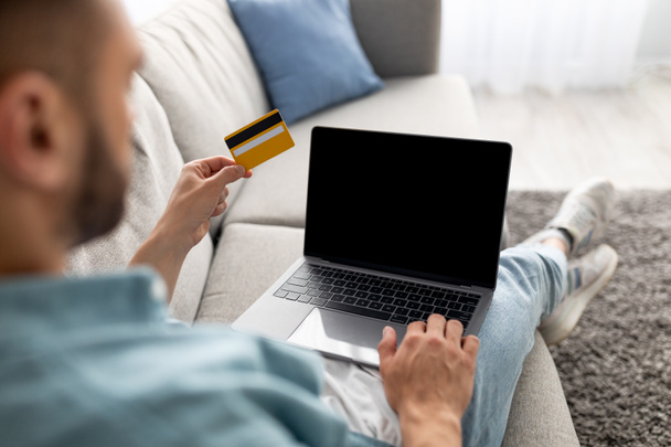 Unbekannter junger Mann sitzt auf Sofa, benutzt Kreditkarte für Online-Einkäufe auf Laptop, Platz für Website-Attrappe - Foto, Bild