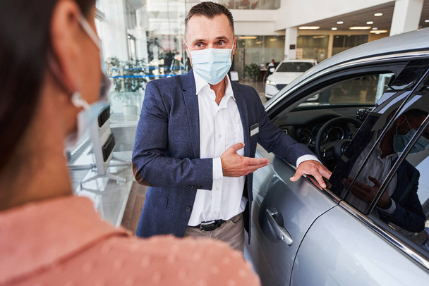 Kadın bir müşteriyle konuşurken tıbbi maske takan satıcı araba seçmeye yardım ediyor. - Fotoğraf, Görsel