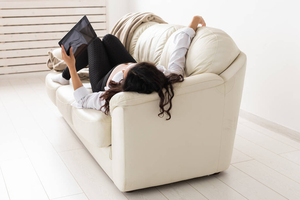 Mujer embarazada latina usando tableta mientras está acostada en el sofá en casa. Embarazo e información para el concepto de paternidad. - Foto, imagen