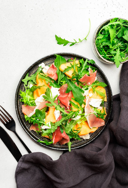 Melon salad with cantaloupe, prosciutto, goat cheese and arugula on white background, top view, copy space - Foto, Bild