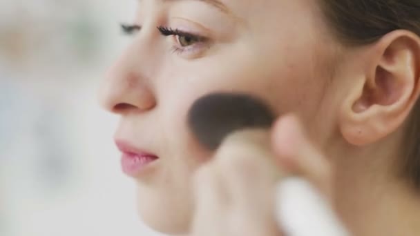 Una joven usa un cepillo de maquillaje para dibujar rubor en sus mejillas. Primer plano - Metraje, vídeo