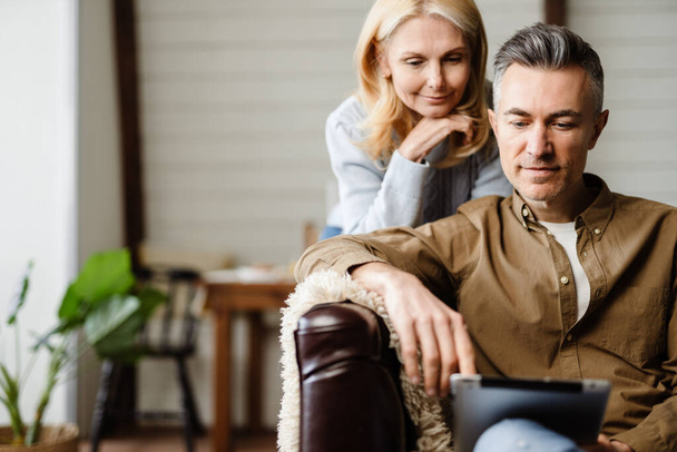 Smiling middle aged white couple relaxing on a couch at home, using tablet device - Foto, Bild
