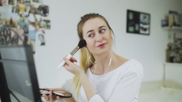 Une jeune femme est assise à une table devant un miroir, écoutant de la musique avec des écouteurs et faisant du maquillage - Séquence, vidéo