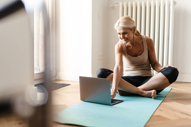 Donna matura bionda che utilizza il computer portatile durante la pratica dello yoga a casa - Foto, immagini