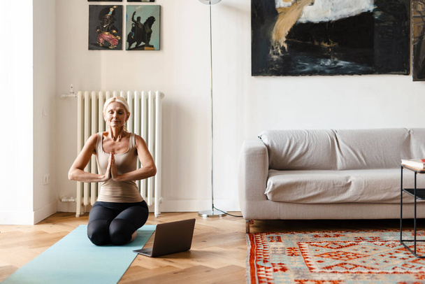 Blonde mature woman using laptop during yoga practice at home - 写真・画像