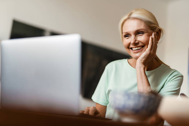 Blonde mature woman smiling and working with laptop while having lunch at home - Foto, Imagen