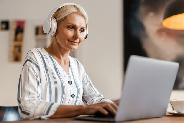 Blonde mature woman in headphones smiling while working with laptop at home - Fotografie, Obrázek