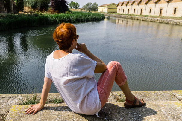 Πίσω όψη μιας γυναίκας με κόκκινα μαλλιά και λευκό πουκάμισο, χαλαρώνοντας στην όχθη ενός ποταμού - Φωτογραφία, εικόνα