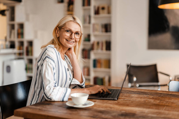 Blonde mature woman in eyeglasses smiling while working with laptop at home - Foto, immagini