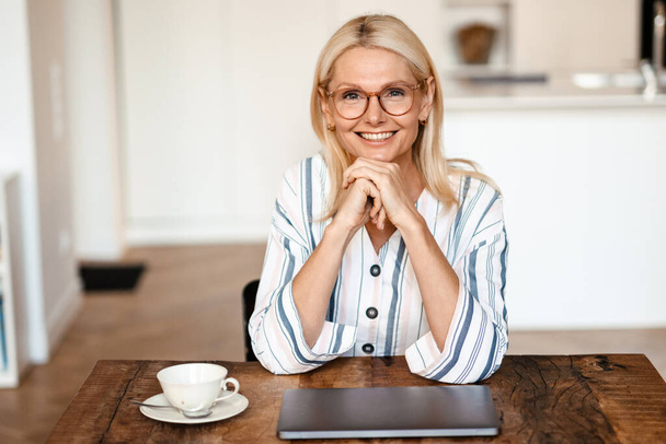 Blonde mature woman wearing eyeglasses smiling while sitting at table in home - 写真・画像