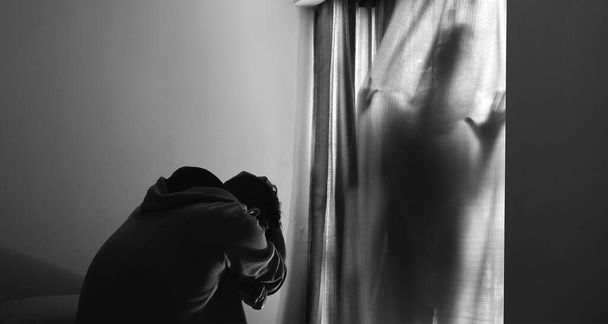 Eine Horizontaufnahme eines verängstigten Mannes, der auf dem Bett sitzt und Angst vor einem Geist hinter den Vorhängen hat - Foto, Bild