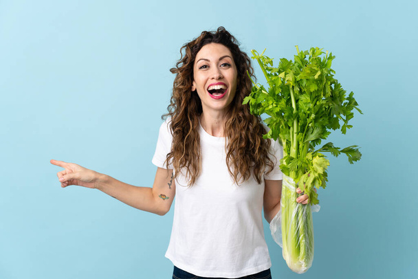 Young woman holding a celery isolated on blue background surprised and pointing finger to the side - Foto, Bild