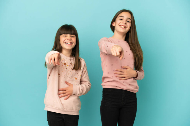 Kleine zusjes meisjes geïsoleerd op blauwe achtergrond wijzend met de vinger naar iemand en lachen veel - Foto, afbeelding