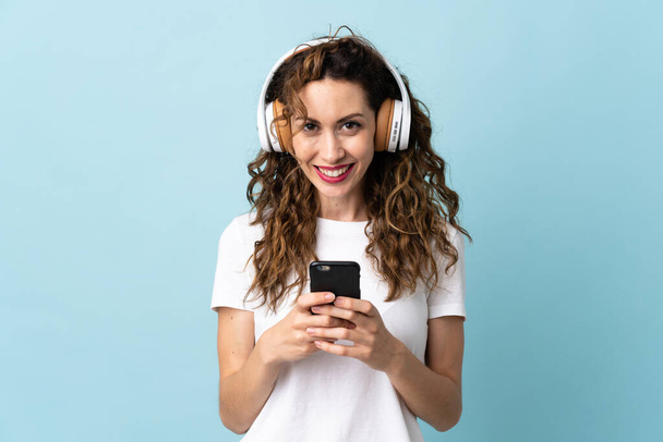 Junge kaukasische Frau isoliert auf blauem Hintergrund Musik hören mit einem Handy und Blick nach vorne - Foto, Bild