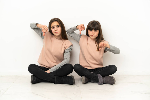 Kleine zusjes zitten op de vloer geïsoleerd op witte achtergrond met duim naar beneden met beide handen - Foto, afbeelding