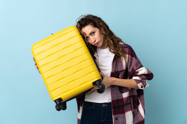 Young caucasian woman isolated on blue background in vacation with travel suitcase and unhappy - Photo, Image