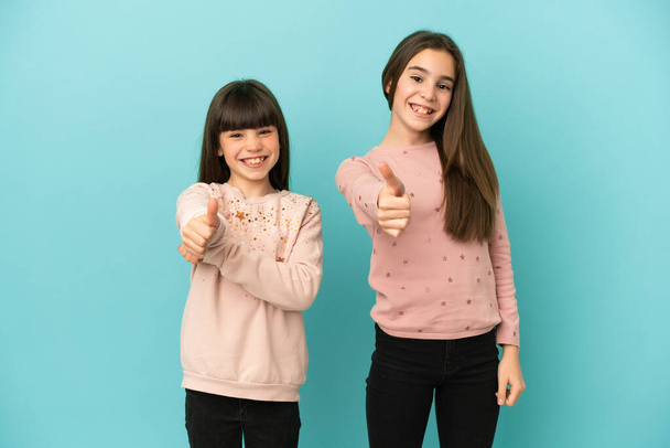 Kleine zusjes meisjes geïsoleerd op blauwe achtergrond geven een duim omhoog gebaar omdat er iets goeds is gebeurd - Foto, afbeelding