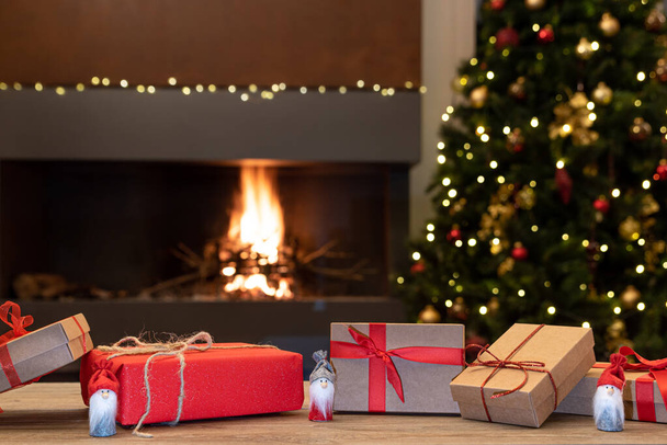 Ambiance de Noël confortable avec des gnomes de Noël et des cadeaux sur une table en bois au premier plan. En arrière-plan brouillé la cheminée avec le feu, le sapin de Noël et les décorations de Noël. Concentration sélective - Photo, image