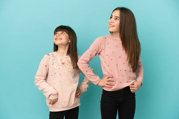 kleine zusjes meisjes geïsoleerd op blauwe achtergrond poseren met armen op heup en lachen - Foto, afbeelding