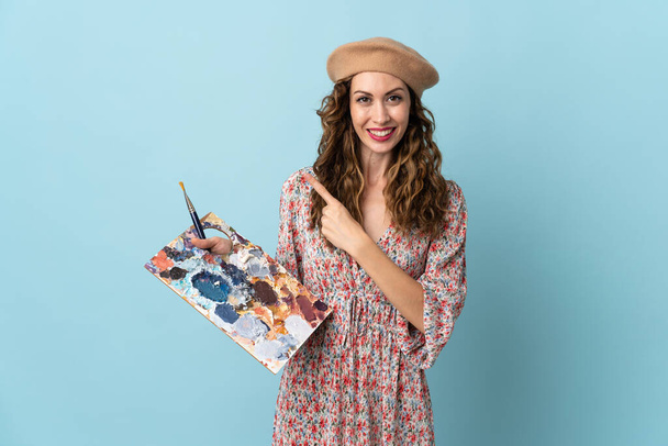 Mladý umělec dívka drží paletu izolované na modrém pozadí ukazuje na stranu prezentovat produkt - Fotografie, Obrázek