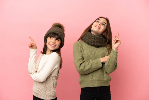 Kleine zusjes dragen een winter kleren geïsoleerd op roze achtergrond wijzen op een geweldig idee en kijken omhoog - Foto, afbeelding