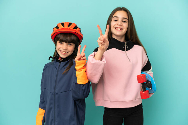 Hermanitas practicando ciclismo y patinadora aisladas sobre fondo azul sonriendo y mostrando señal de victoria - Foto, Imagen