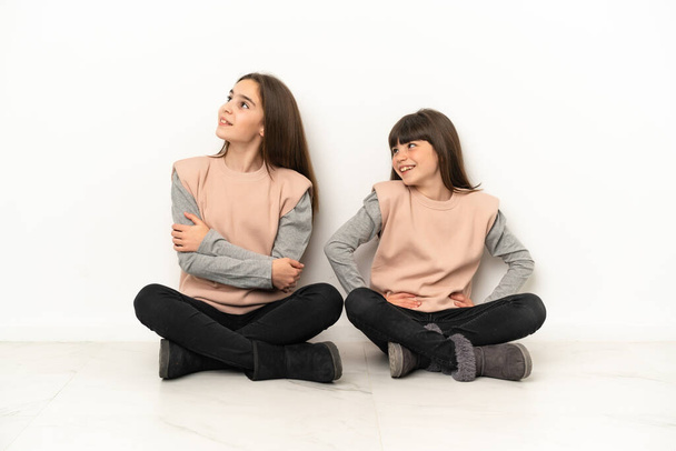 Kleine zusjes zitten op de vloer geïsoleerd op witte achtergrond poseren met armen op heup en lachen - Foto, afbeelding