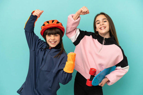 Kleine Schwestern üben Radfahren und Skater isoliert auf blauem Hintergrund feiern einen Sieg - Foto, Bild