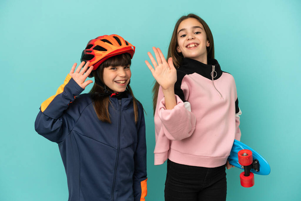 Little sisters practicing cycling and skater isolated on blue background saluting with hand with happy expression - Foto, Imagem