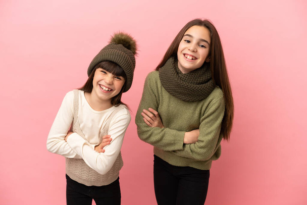 Kleine zusjes dragen een winter kleren geïsoleerd op roze achtergrond houden de armen gekruist terwijl ze glimlachen - Foto, afbeelding