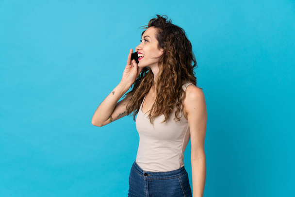 Young caucasian woman isolated on blue background keeping a conversation with the mobile phone - Фото, изображение