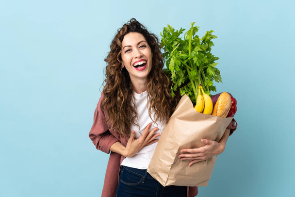 Mujer joven sosteniendo una bolsa de la compra de comestibles aislado sobre fondo azul sonriendo mucho - Foto, imagen