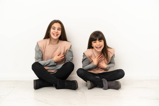 Hermanitas sentadas en el suelo aisladas sobre fondo blanco sonriendo mucho mientras ponen las manos en el pecho - Foto, imagen
