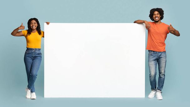 Ευτυχισμένο αφρο-αμερικανικό ζευγάρι στέκεται δίπλα σε κενό οριζόντιο πίνακα για διαφήμιση - Φωτογραφία, εικόνα
