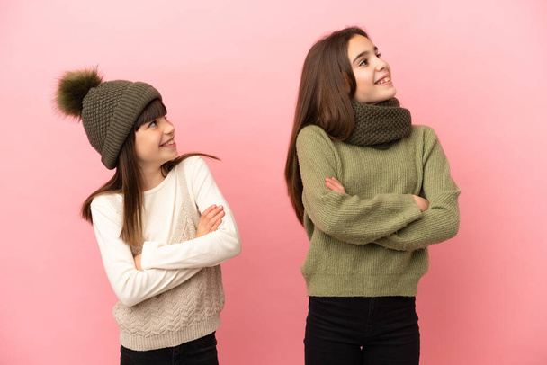 Hermanitas vistiendo una ropa de invierno aisladas sobre fondo rosa mirando hacia arriba mientras sonríen - Foto, imagen
