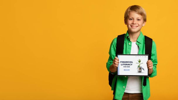 Веселый мальчик демонстрирует планшетный компьютер с веб-сайтом школы Financial Literacy for Kids на оранжевом фоне - Фото, изображение
