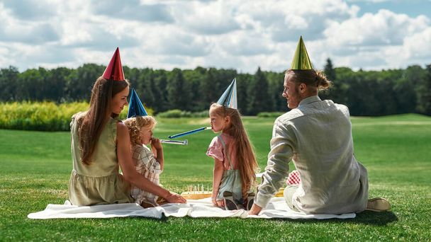 Visão traseira da família feliz vestindo chapéus de festa, celebrando o aniversário das crianças, fazendo piquenique no parque em um dia de verão. Crianças assobiando assobios - Foto, Imagem