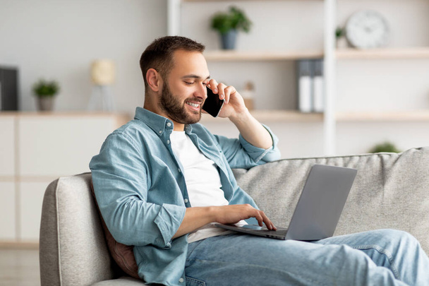 Attractive young guy talking on smartphone and using laptop computer on couch at home office - 写真・画像