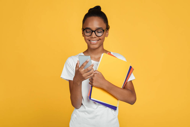 Smiling teenager african american girl pupil in glasses and casual holds books and reads message on smartphone - Photo, Image