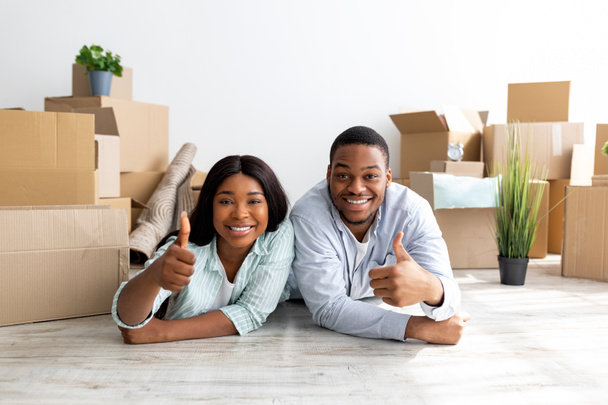 Happy black couple recommending real estate agency, showing thumbs up while lying on floor among cardboard boxes - Foto, imagen