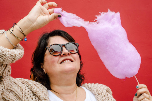 Mulher rasga um pedaço de algodão doce lilás para comê-lo, usa óculos escuros e está em um fundo vermelho - Foto, Imagem