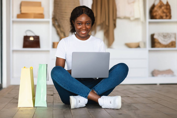 African Lady Shopping con computadora portátil sentada cerca de bolsas de compras en interiores - Foto, Imagen